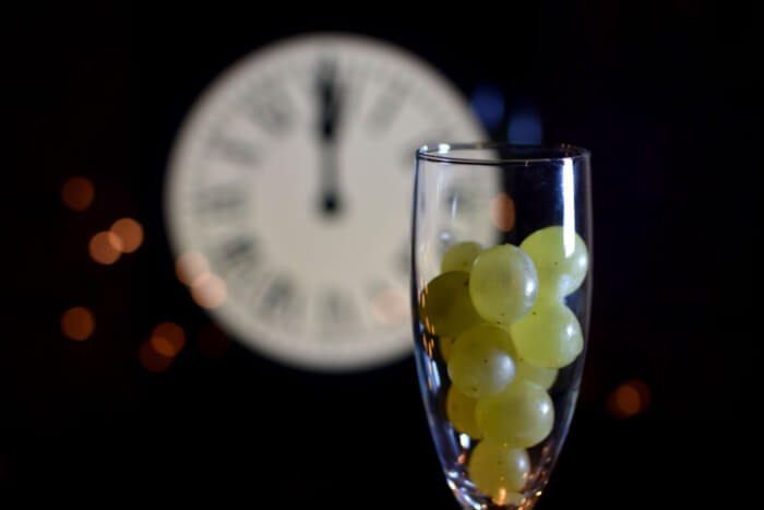 Uma taça com 12 uvas é destaque, ao fundo, o relógio batendo meia-noite.