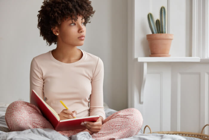 Mulher negra sentada na cama com um caderno em mãos. Pensando em como organizar as tarefas do dia a dia. 