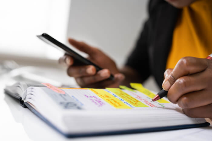 Mulher negra utilizando o caderno e o celular para organizar suas tarefas em uma agenda.