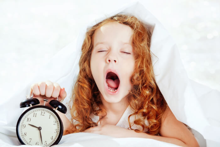 Menina bocejando no primeiro dia de volta às aulas com um despertador ao lado.