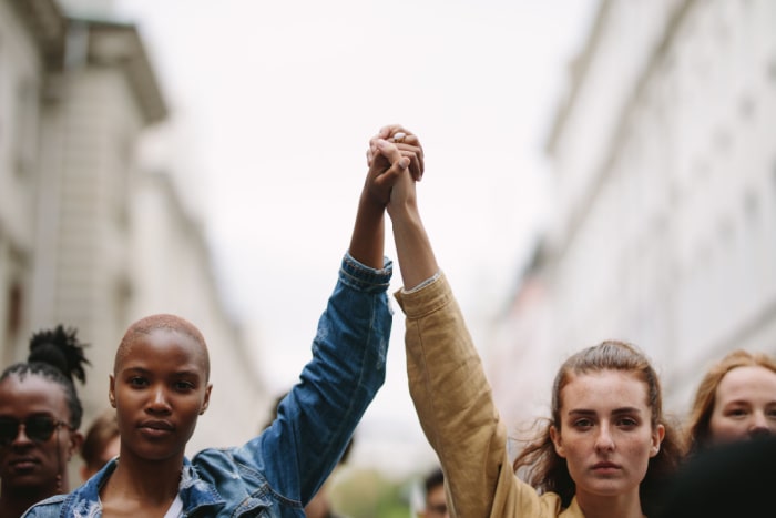 Duas mulheres de mãos dadas e para o alto em meio a uma marcha pela equidade de gênero.